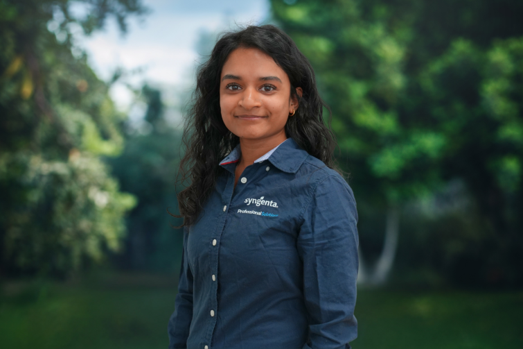 Smiling woman in a blue button-up shirt outdoors with blurred greenery background